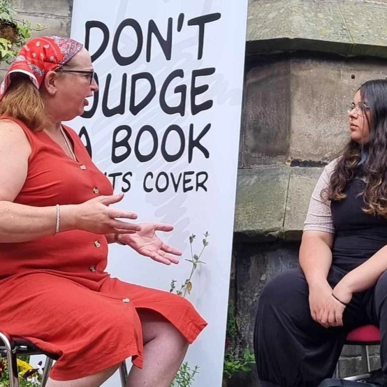 Our book of the month with her reader at an event in Scotland. They are sitting outside in front of a Human Library banner that says "dont judge a book by its cover"