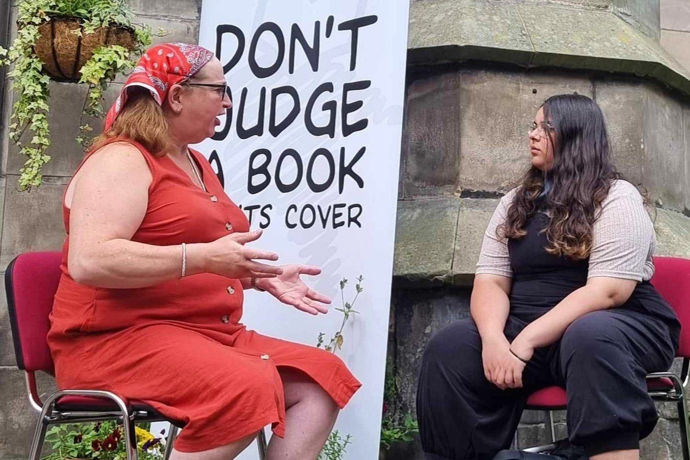 Our book of the month with her reader at an event in Scotland. They are sitting outside in front of a Human Library banner that says "dont judge a book by its cover"