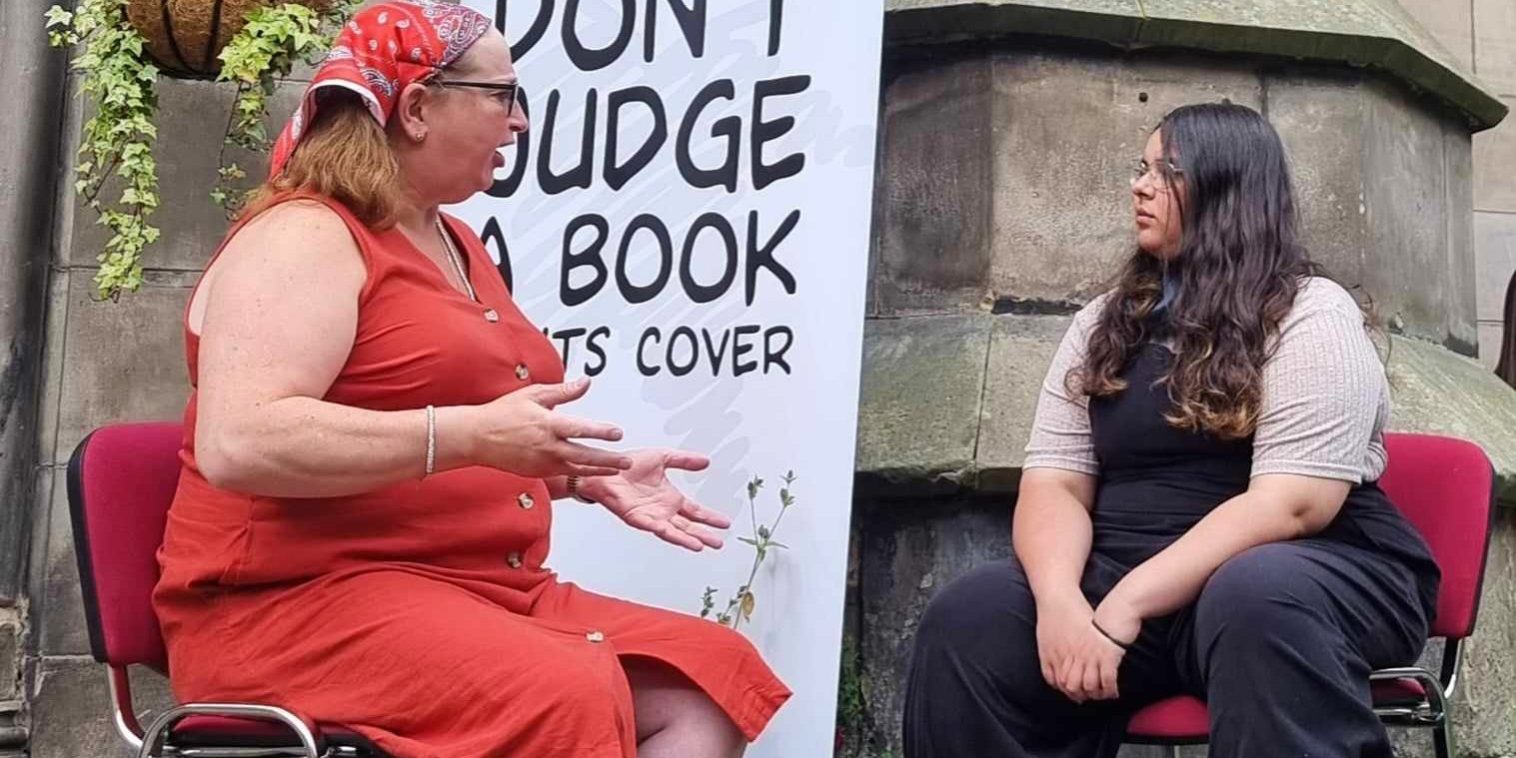 Rachel Book of the Month Our book of the month with her reader at an event in Scotland. They are sitting outside in front of a Human Library banner that says "dont judge a book by its cover"