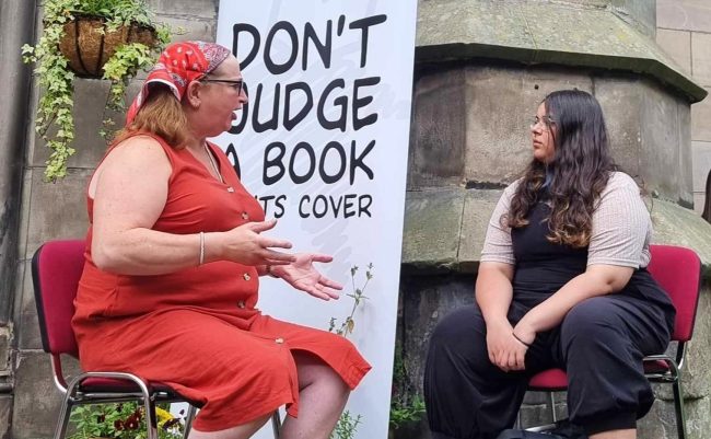 Our book of the month with her reader at an event in Scotland. They are sitting outside in front of a Human Library banner that says "dont judge a book by its cover"