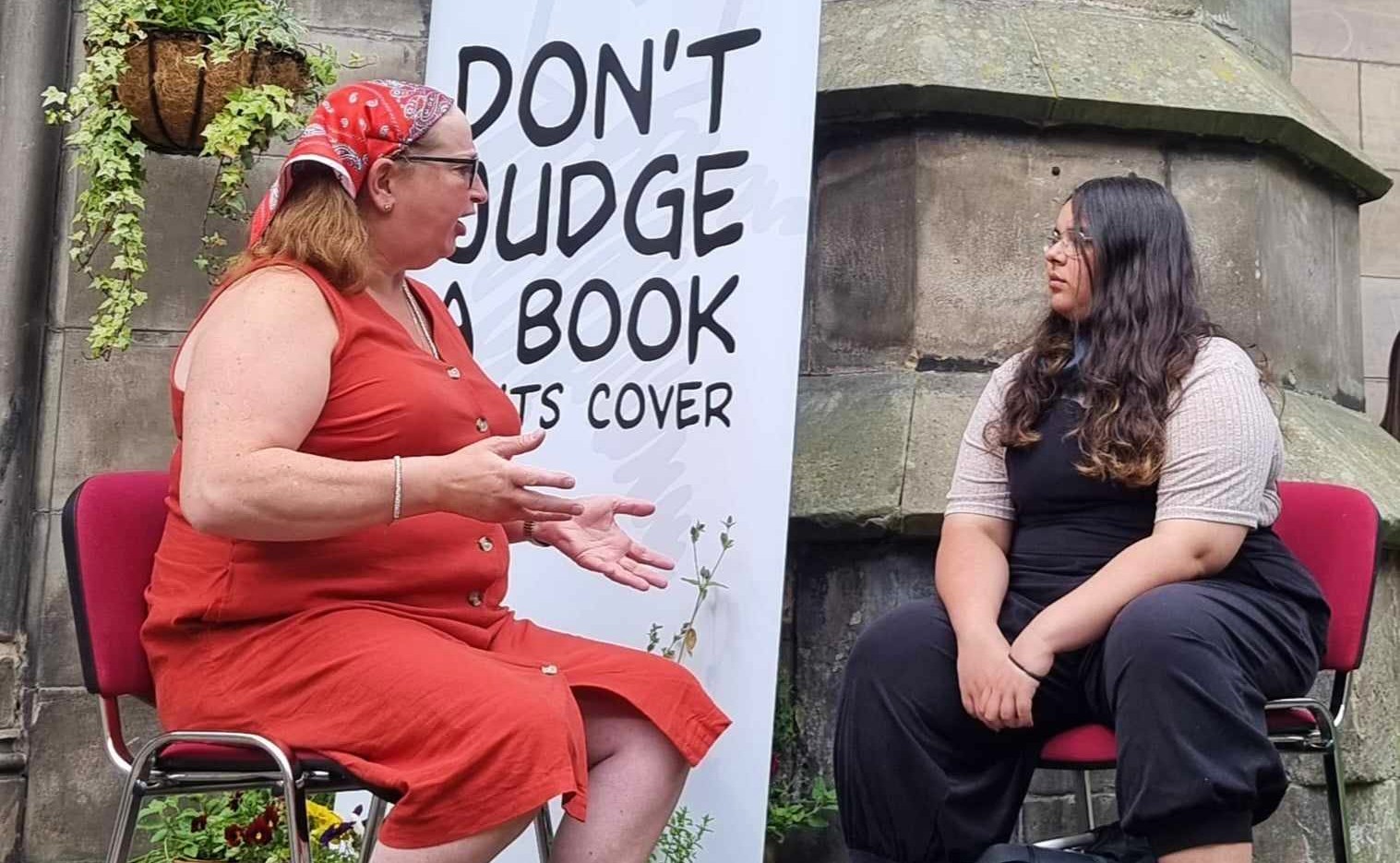 Our book of the month with her reader at an event in Scotland. They are sitting outside in front of a Human Library banner that says "dont judge a book by its cover"