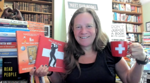Katy holding a mug featuring the Swiss flag and a book titled How to be Swiss, clearly curious about what that entails.